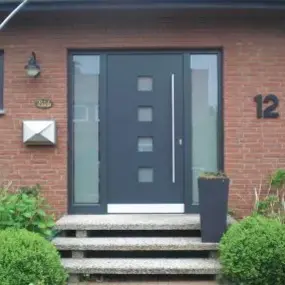 Moderne Haustür aus Glas und Metall in einem roten Klinkerhaus mit Hausnummer 12.