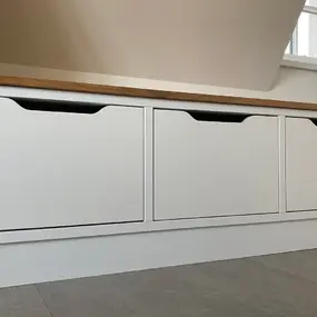 Weiße Sitzbank mit drei Schubladen und Holzplatte unter einer Dachschräge im Raum.