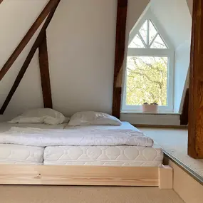 Gemütliches Schlafzimmer im Dachgeschoss mit Holzbalken, Doppelbett und großem Fenster.